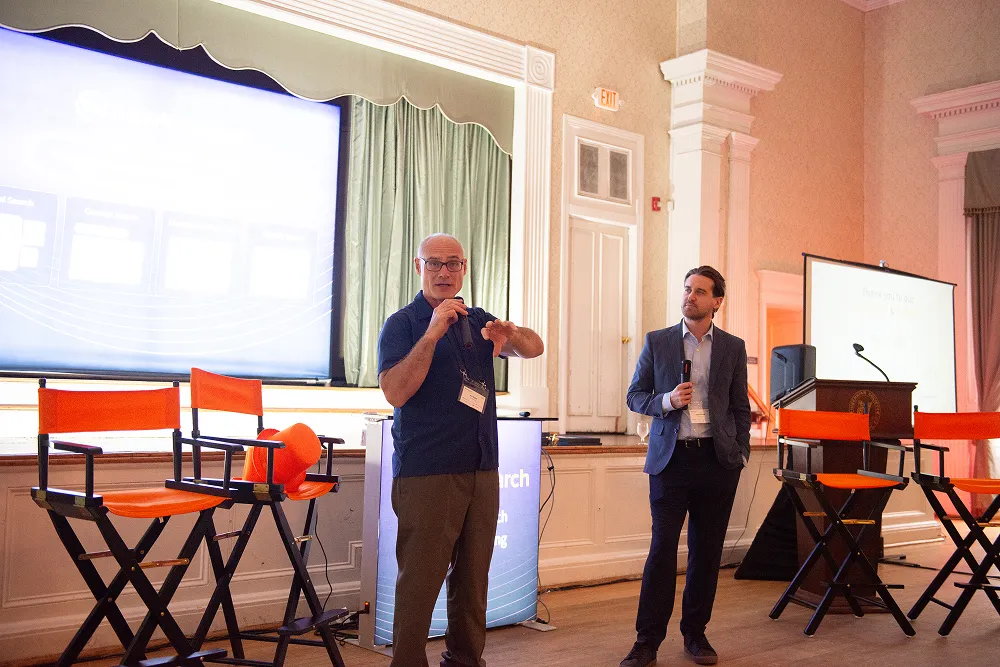 Ari Kahn and Kyle Mitzner lead a discussion on stage during the HawkSearch Customer Summit 25, with orange chairs and branded signage in the background.
