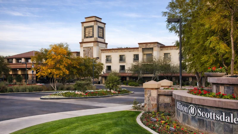Exterior view of Hilton Scottsdale Resort and Villas with landscaped roundabout, green lawn, and trees in front.