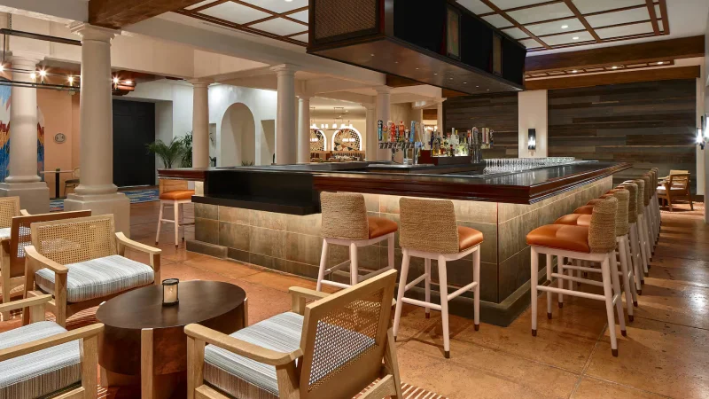 Modern bar area with a large square counter surrounded by wicker and leather bar stools, wooden chairs with cushions, and warm ambient lighting.