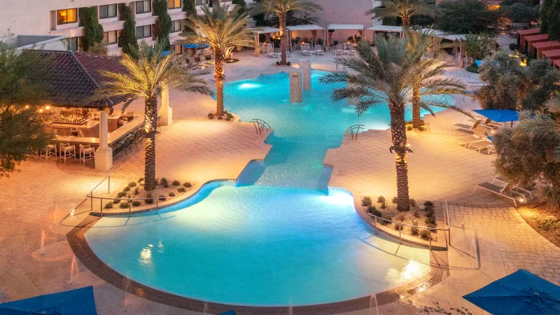 Illuminated outdoor swimming pool area at dusk with palm trees, lounge chairs, and a shaded bar area.
