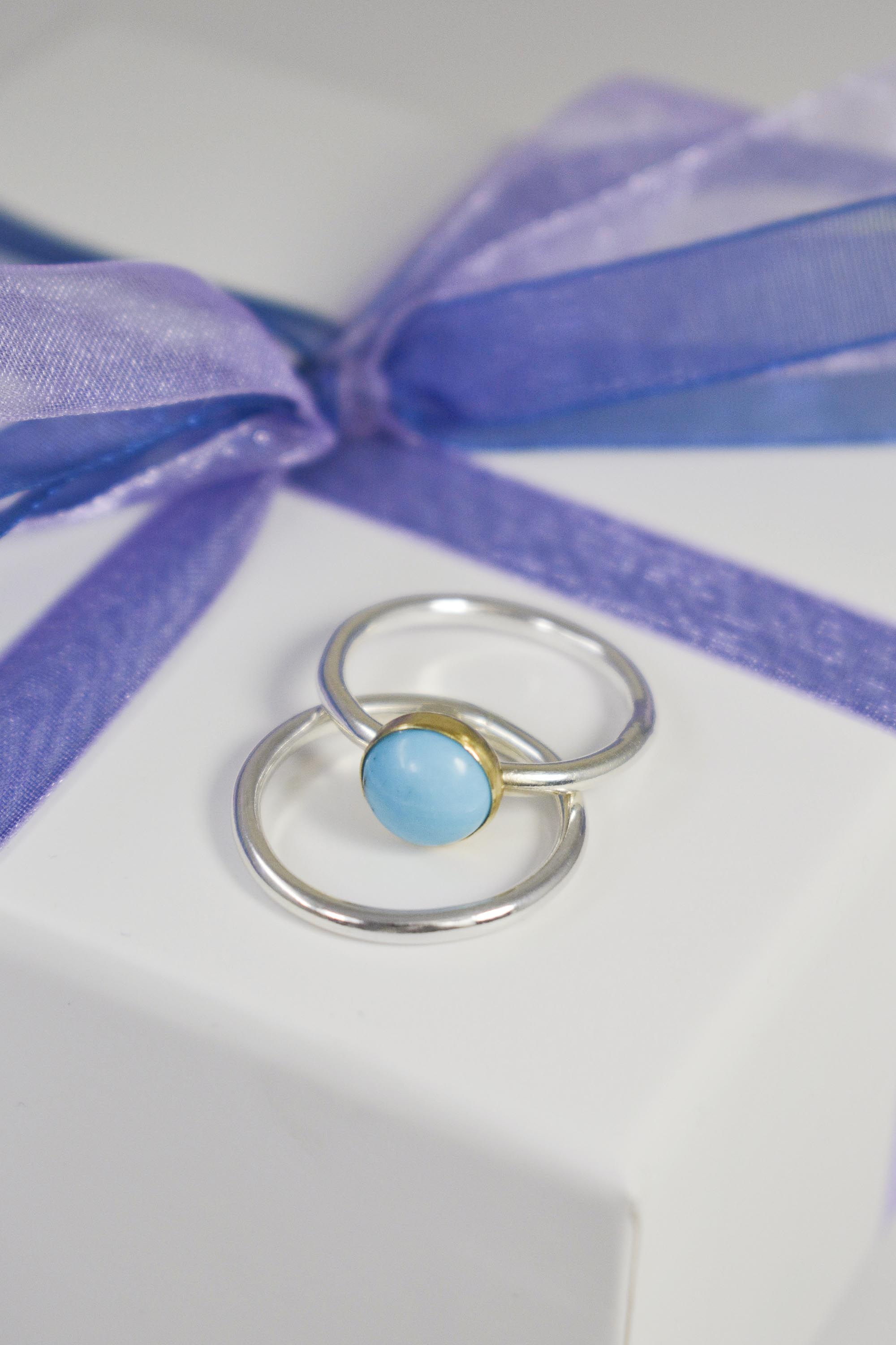 turquoise gemstone ring in gold bezel with silver ring shank and silver stacking ring resting on white jewellery box tied with navy ribbon