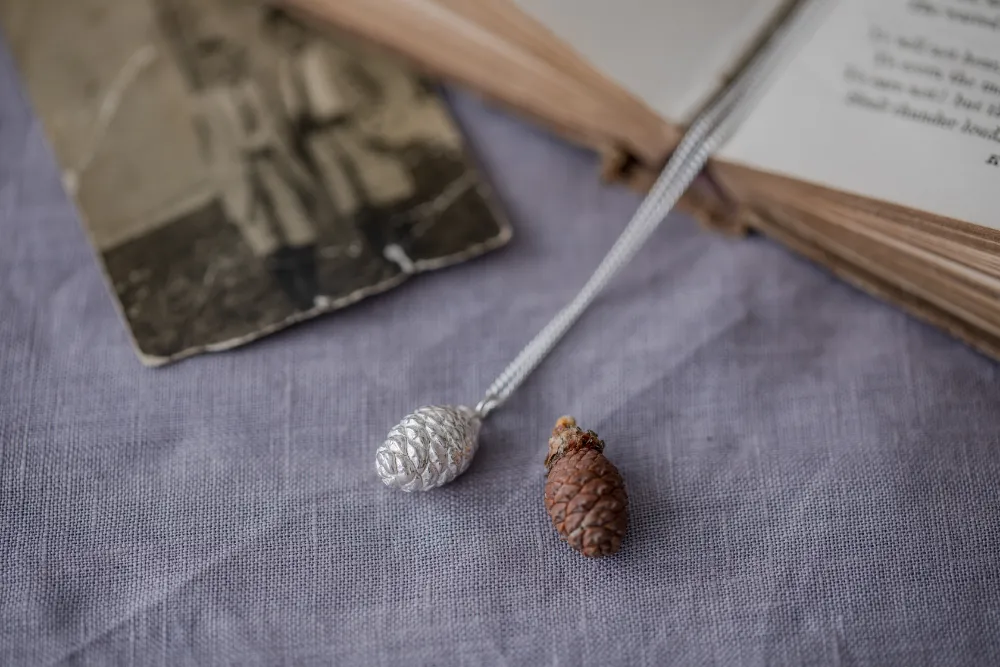 silver pine cone necklace with original pine cone on pale purple linen tablecloth and blurred image of photograph of boys resting on open book