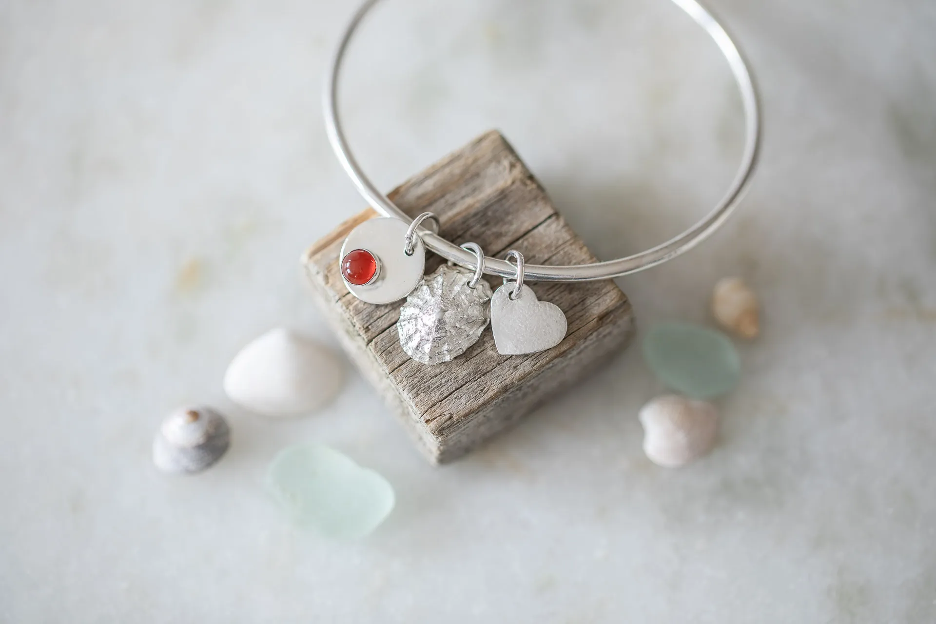 bangle with carnelian charm, limpet shell and heart charms on piece of driftwood with shells in the foreground on marble background