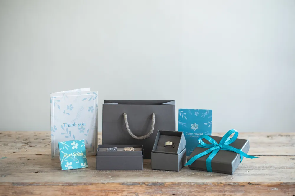 CHJ packaging for mens jewellery with grey jewellery boxes tied with ribbon, care card, polishing cloth and thank you card on pale wooden table