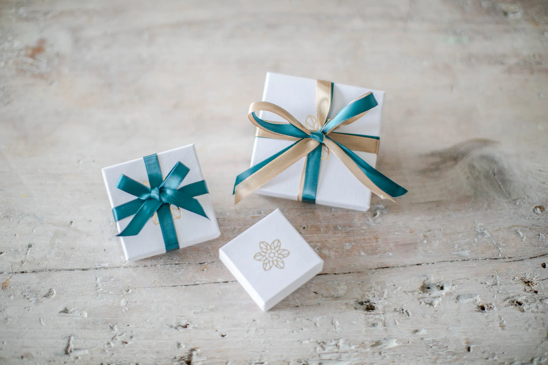 3 white jewellery boxes with gold flower logo tied with teal ribbon resting on pale wooden background