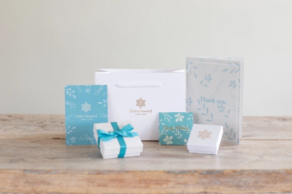 CHJ packaging including white jewellery box with gold embossed logo tied with ribbon, care card, thank you card, polishing cloth and white jewellery bag