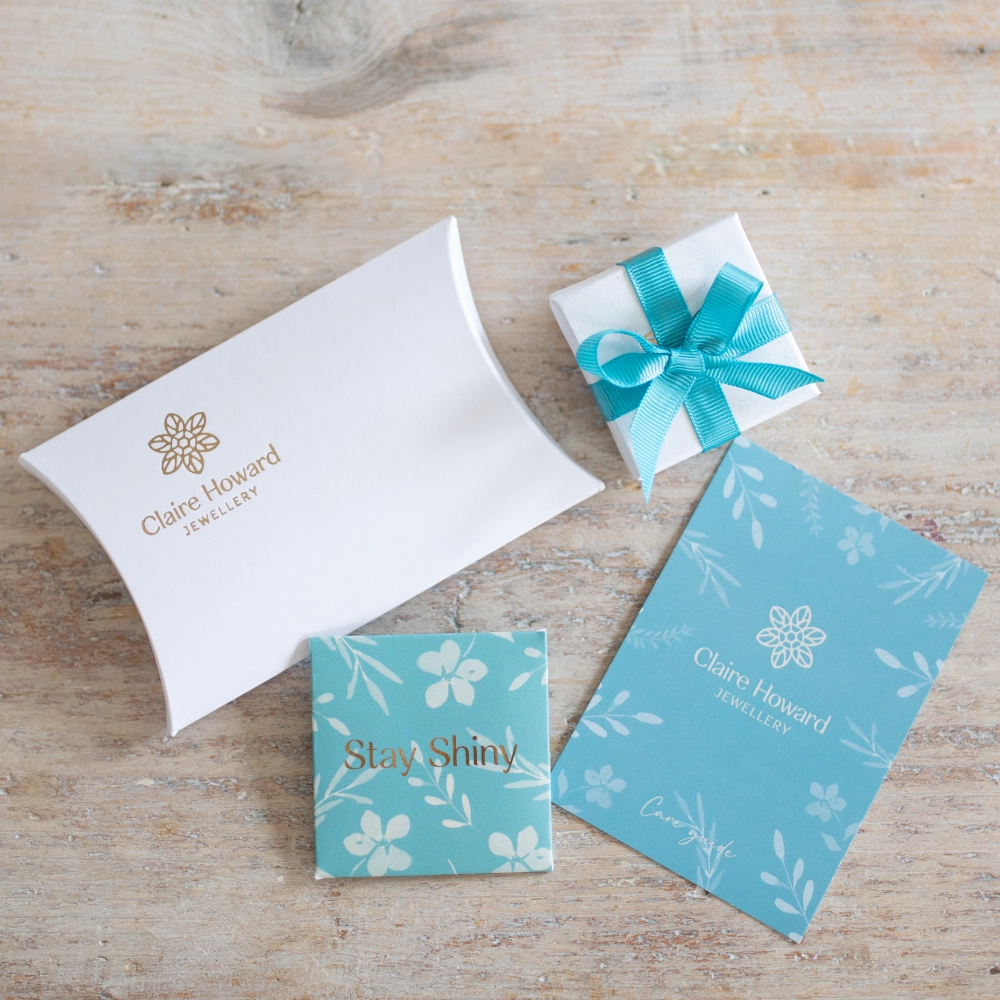 CHj packaging including white pillow box with gold embossed logo, jewellery box tied with blue ribbon, cafre card and polishing cloth