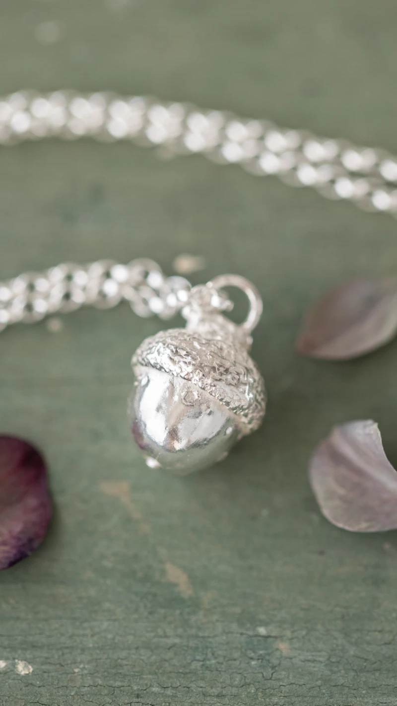 A sliver acorn necklace resting on a green table with flower petals around it