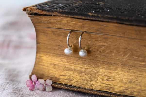 silver and ivory pearl hoop earrings held in gold pages of vintage book on pink ablecloth with small flowers in the foreground
