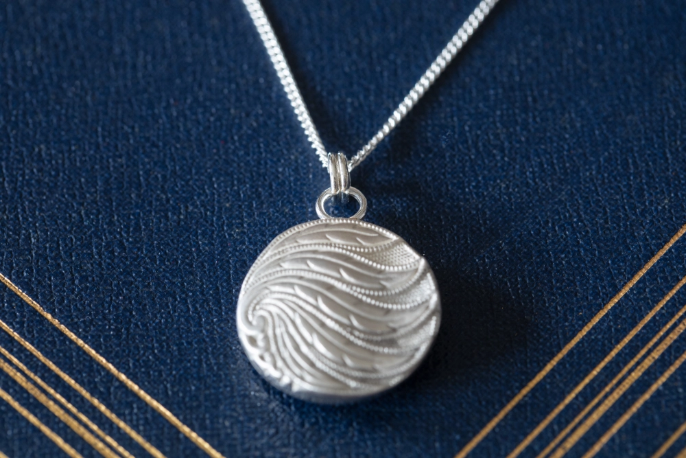 Whispering feather necklace showing wavy feather like lines on front resting on vintage blue book with gold detailing