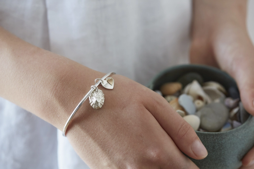 silver limpet shell and heart charm bangle worn by woman in white linen dress holding a bowl of shells