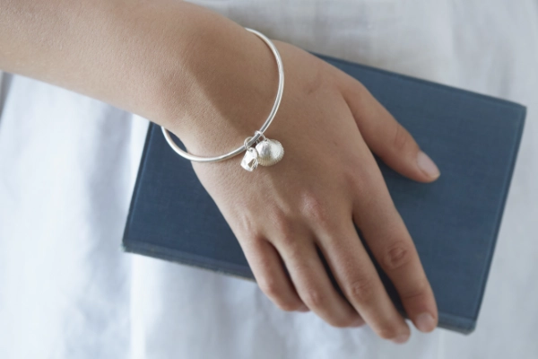 cockle shell and heart bangle worn on wrist of model wearing a white linen dress and holding a vintage blue book