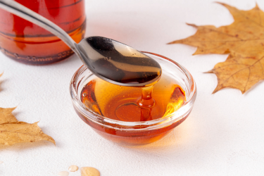 A spoon drizzles amber maple syrup into a glass bowl surrounded by autumn maple leaves.