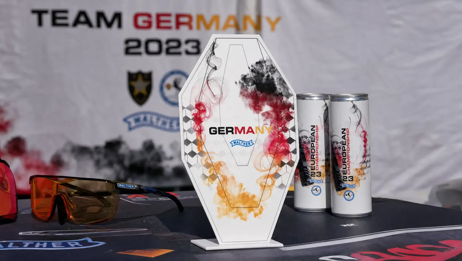 Tischaufsteller „Germany“ und Getränkedosen zur IPSC-Europameisterschaft 2023 vor dem Banner des deutschen Teams