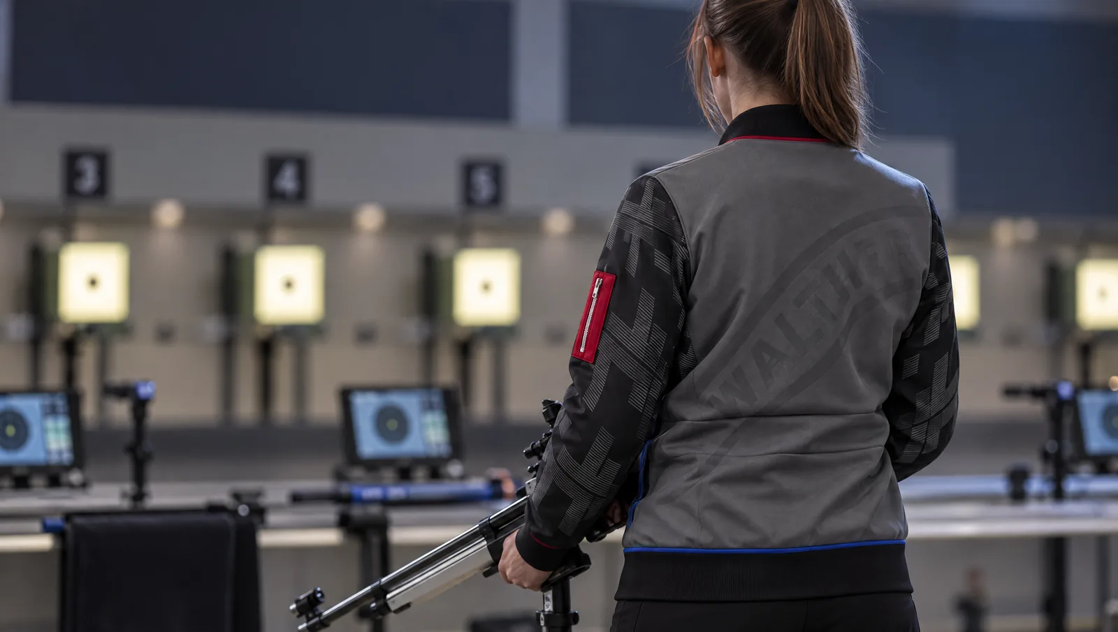 Rückansicht der grauen Schützenjacke von Carl Walther mit großem Walther-Schriftzug, getragen von einer Sportschützin am Schießstand