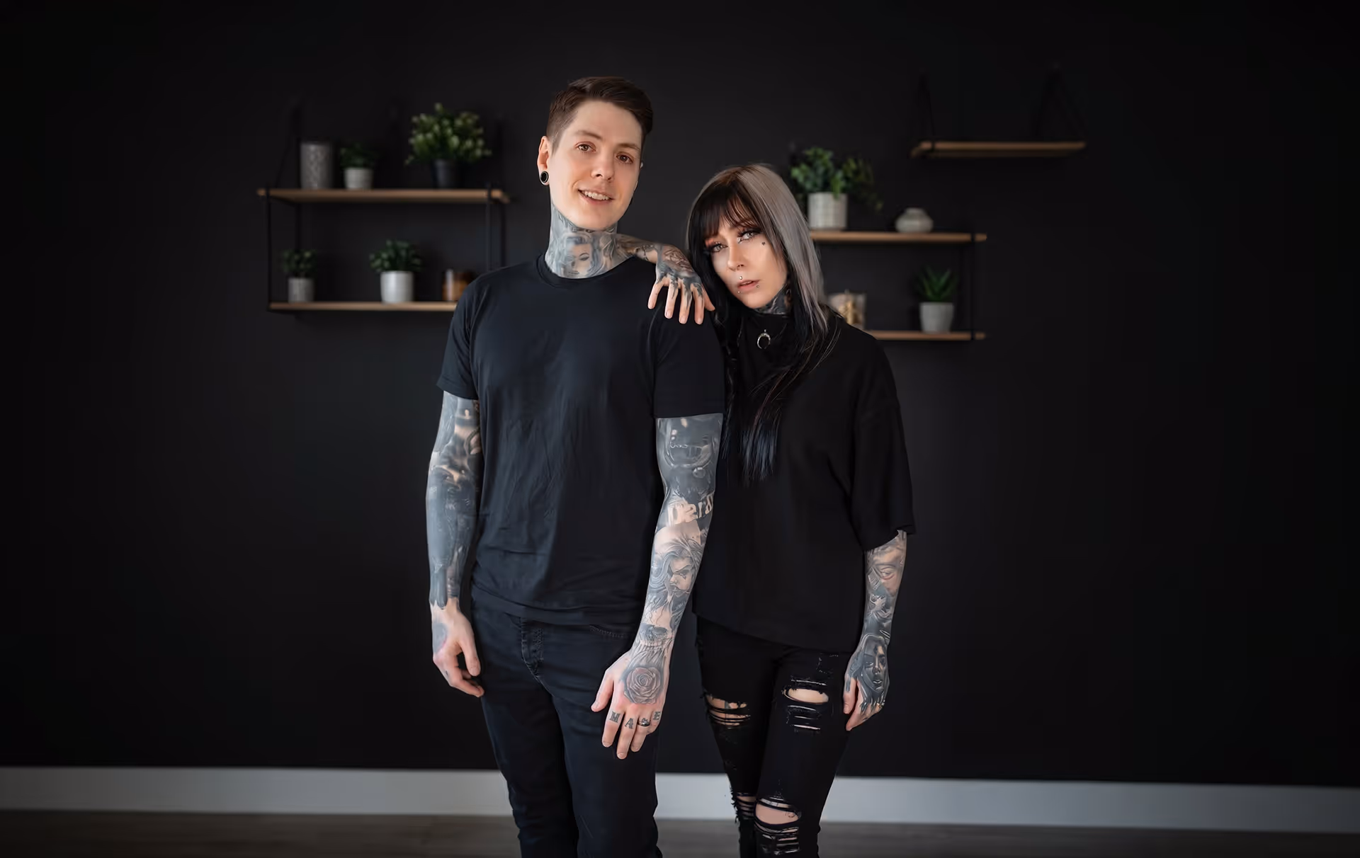 Helen und Stefan, stehend vor einem schwarzen Hintergrund mit Pflanzen auf dem Regal