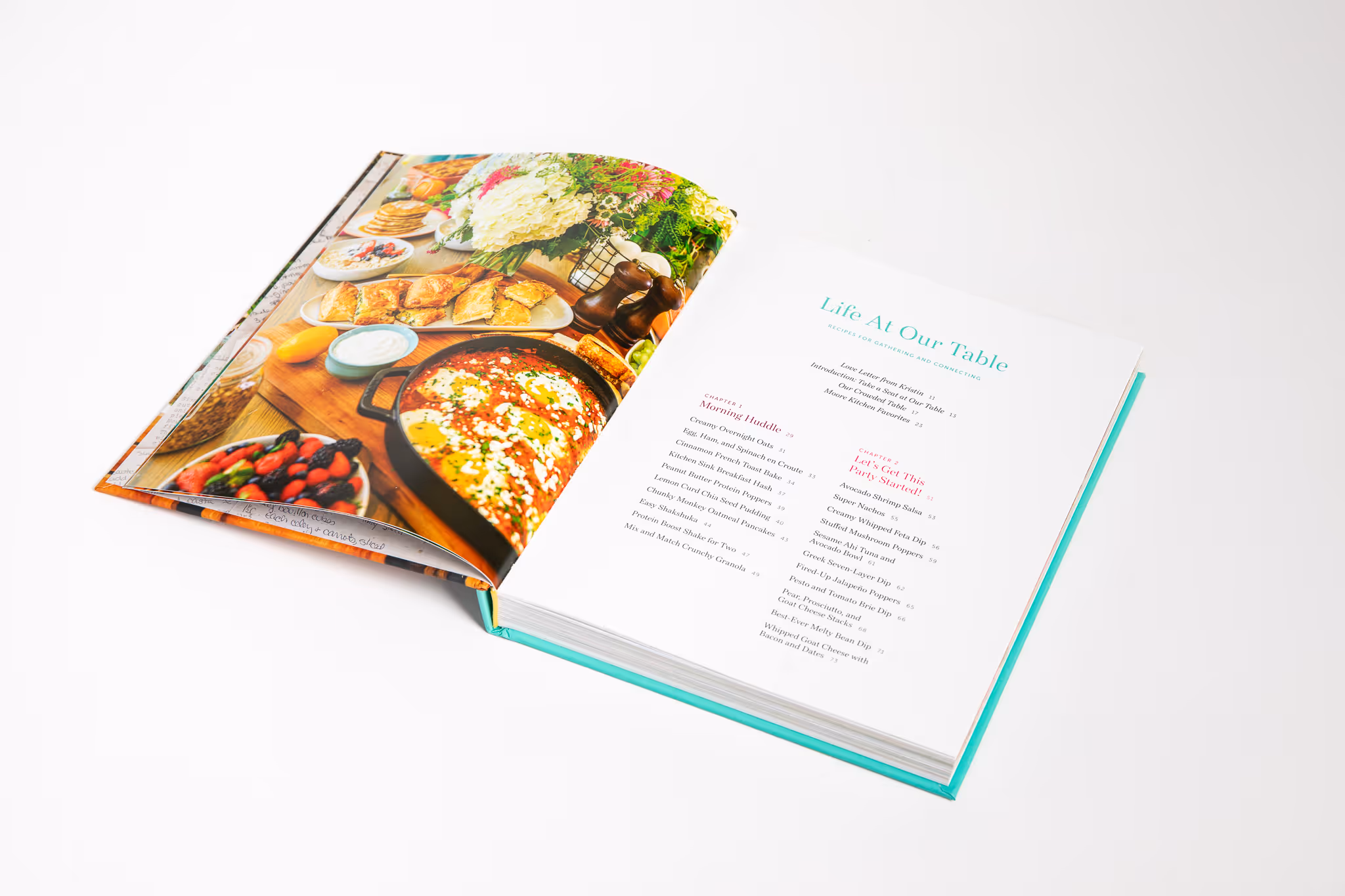 Open cookbook titled 'Life At Our Table' showing a colorful food photo on the left page and a table of contents on the right page.
