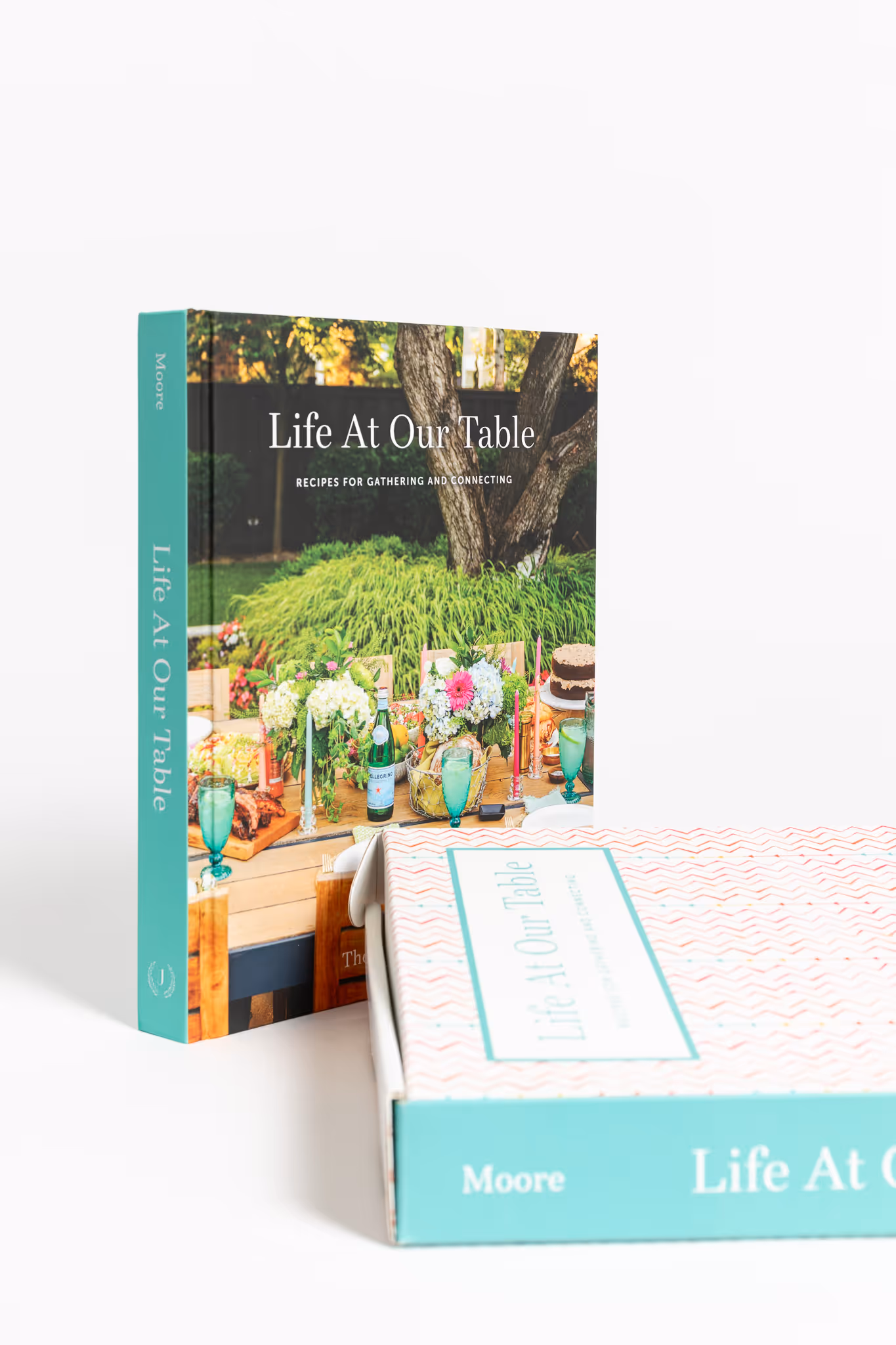 Cookbook titled 'Life At Our Table' with a cover showing a decorated outdoor dining table with flowers, drinks, and food.