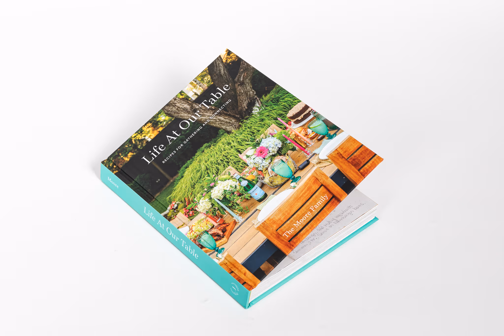 Cookbook titled 'Life At Our Table' featuring an outdoor dining table set with flowers, drinks, and food.