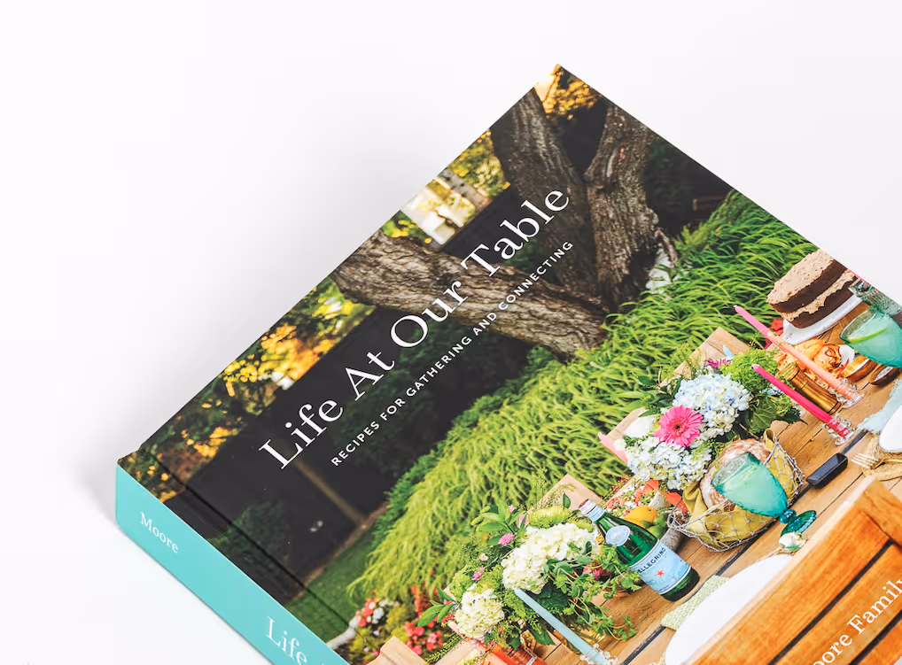 Cookbook titled 'Life At Our Table' with subtitle 'Recipes for Gathering and Connecting' featuring a vibrant outdoor table setting with flowers, drinks, and cake.