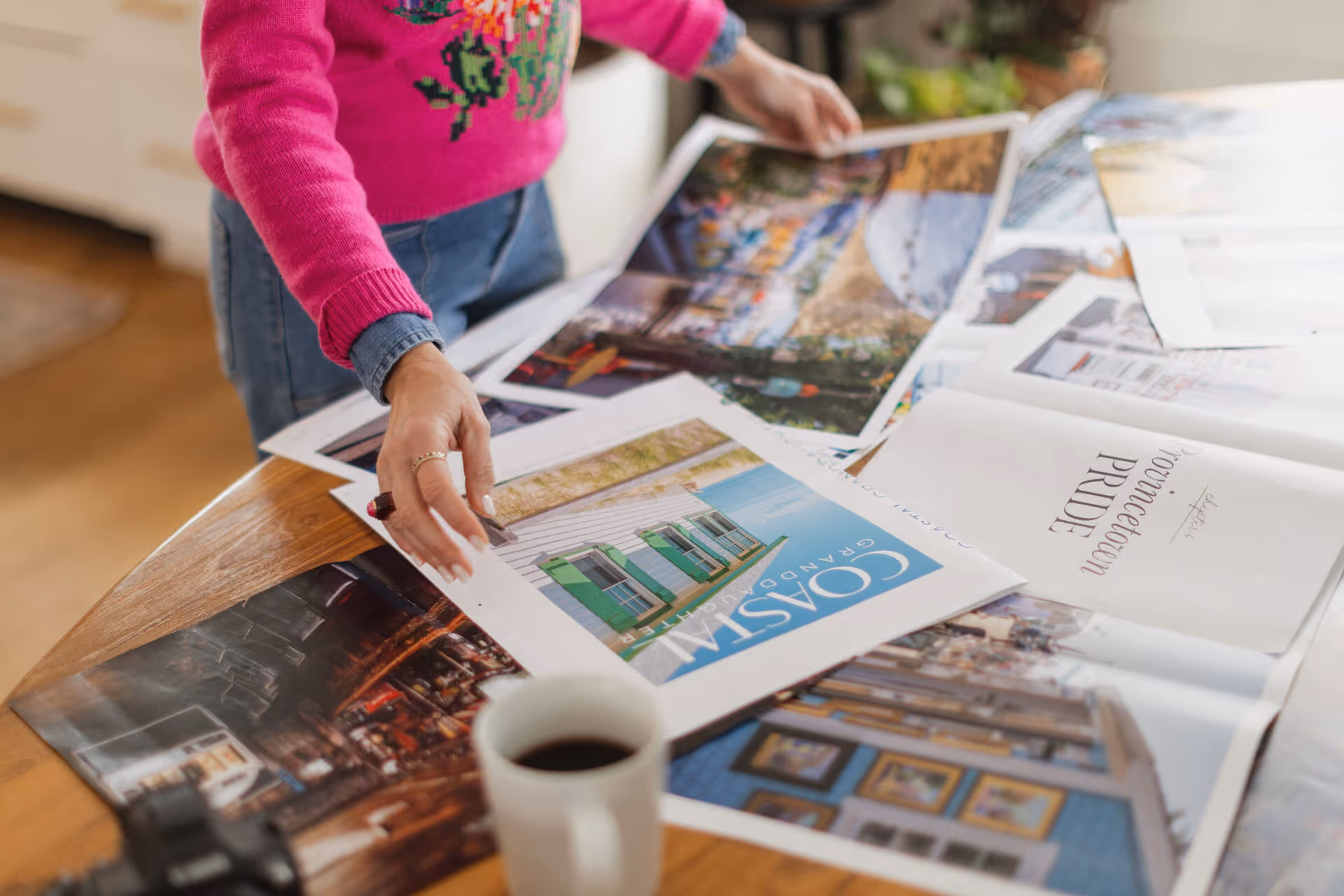 Person in pink sweater and denim shirt arranging colorful travel brochures and a coffee cup on a wooden table.
