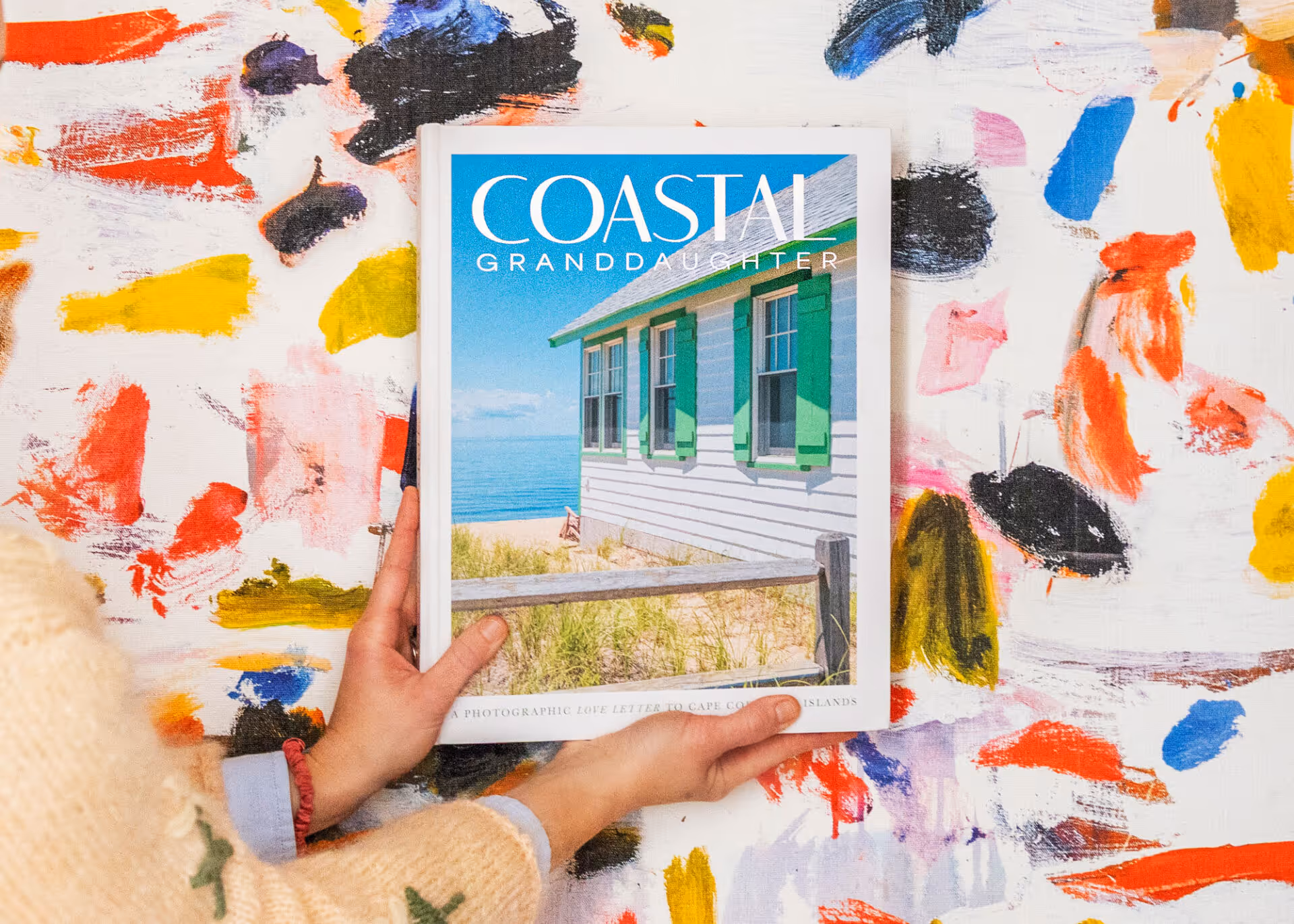 Person holding a book titled 'Coastal Granddaughter' with a cover photo of a white house with green shutters by the beach, against a colorful abstract painted background.