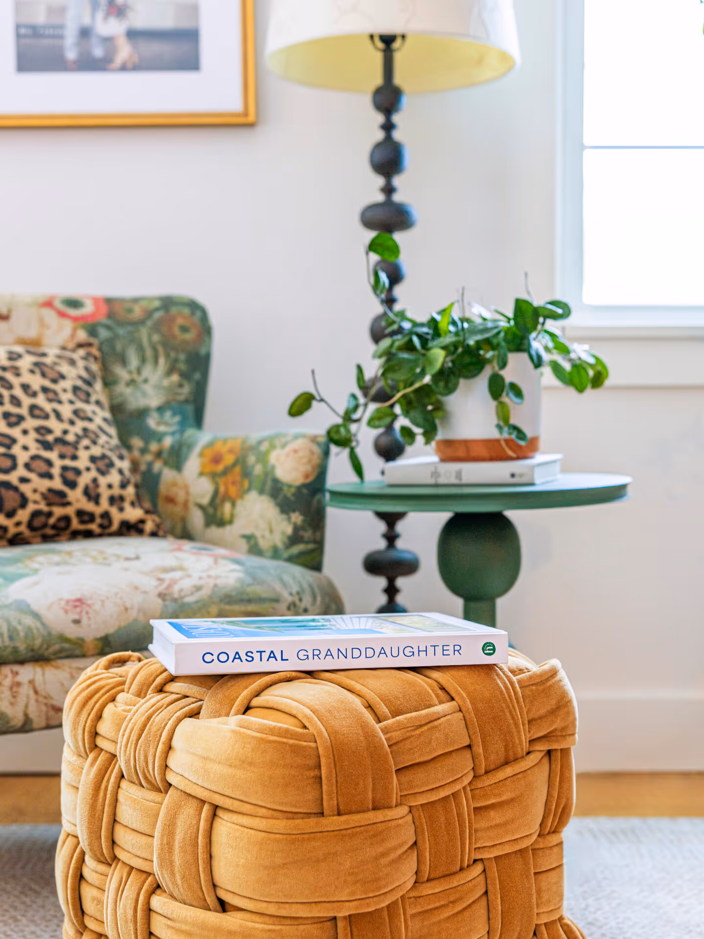 Cozy living room corner with a woven mustard ottoman holding the book 'Coastal Granddaughter,' a floral armchair with leopard print pillow, and a green side table with a potted plant and lamp.