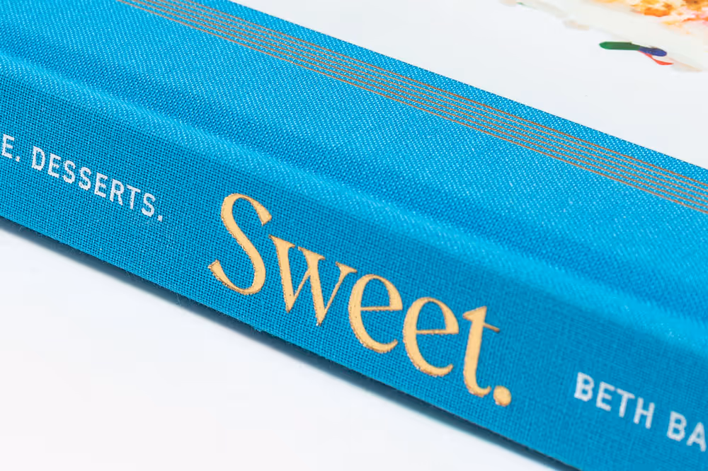 Spine of a turquoise cookbook titled 'Sweet.' by Beth Baumgartner Cook with gold and white foil stamped text.