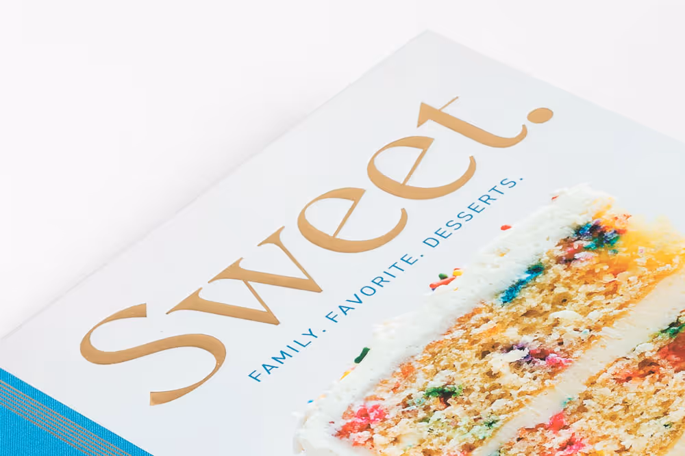 Cookbook titled 'Sweet' with subtitle 'Family. Favorite. Desserts.' and a close-up image of a slice of colorful sprinkle cake with white frosting and gold foil stamping