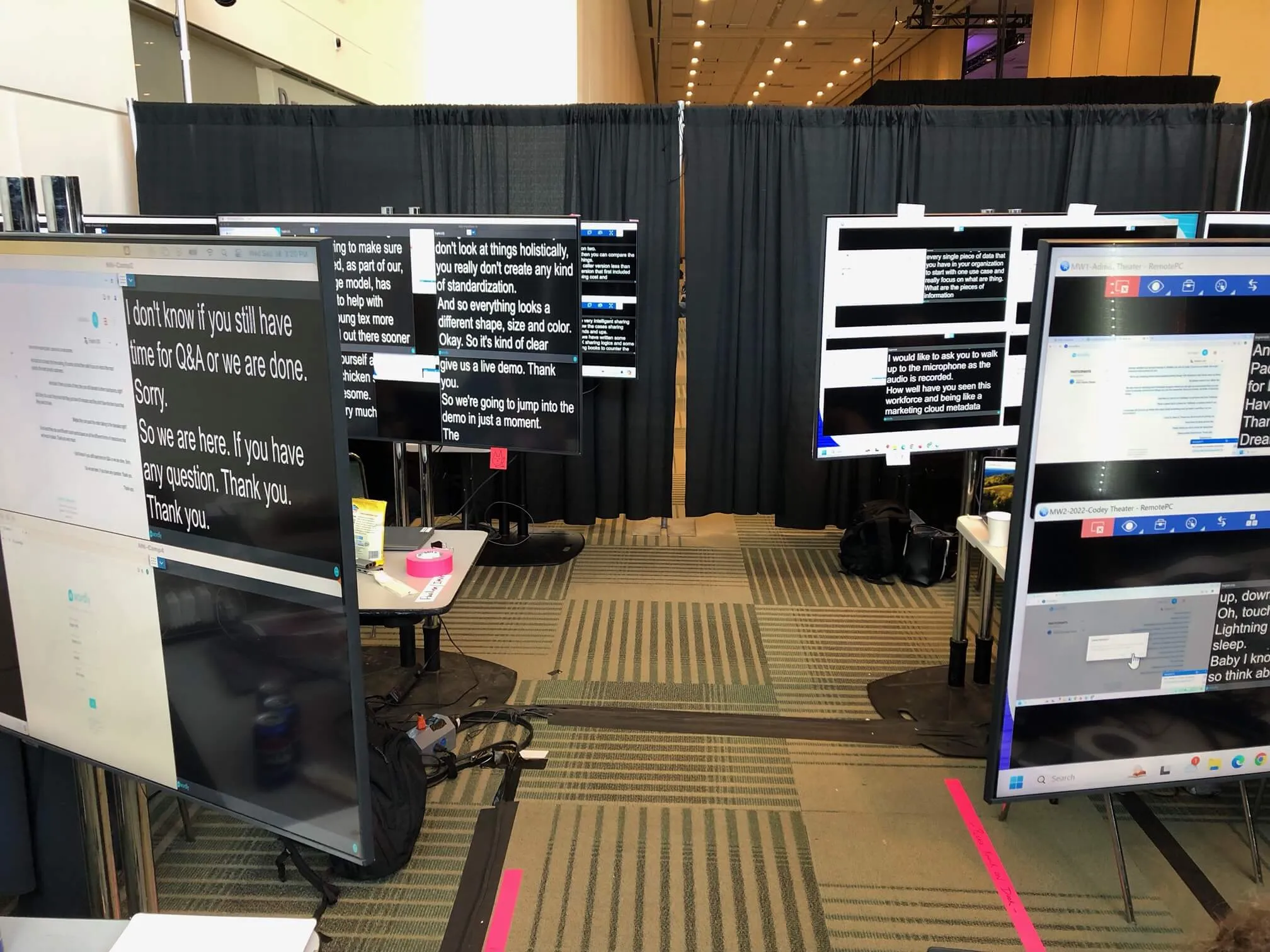 Multiple large monitors display live captions in the Wordly control room behind the scenes at a technology conference. Technicians remotely manage sessions using laptops and software to monitor live AI captioning and translations across event stages.