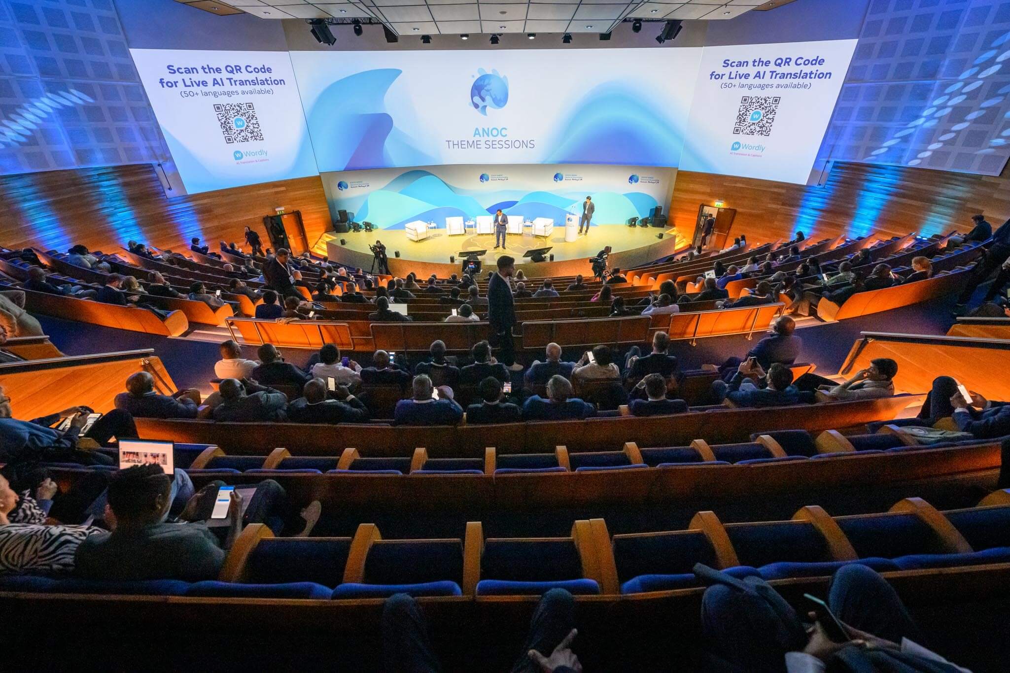 An international conference hall with attendees facing speakers on stage. Two projection screens display a QR code and the message “Scan the QR Code for Live AI Translation” with the Wordly logo