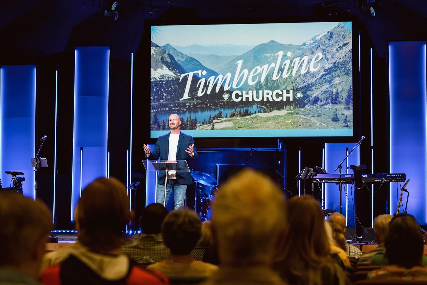 A pastor speaks to the congregation at Timberline Church, standing on stage with a large screen behind him displaying the Timberline Church logo and a mountain landscape