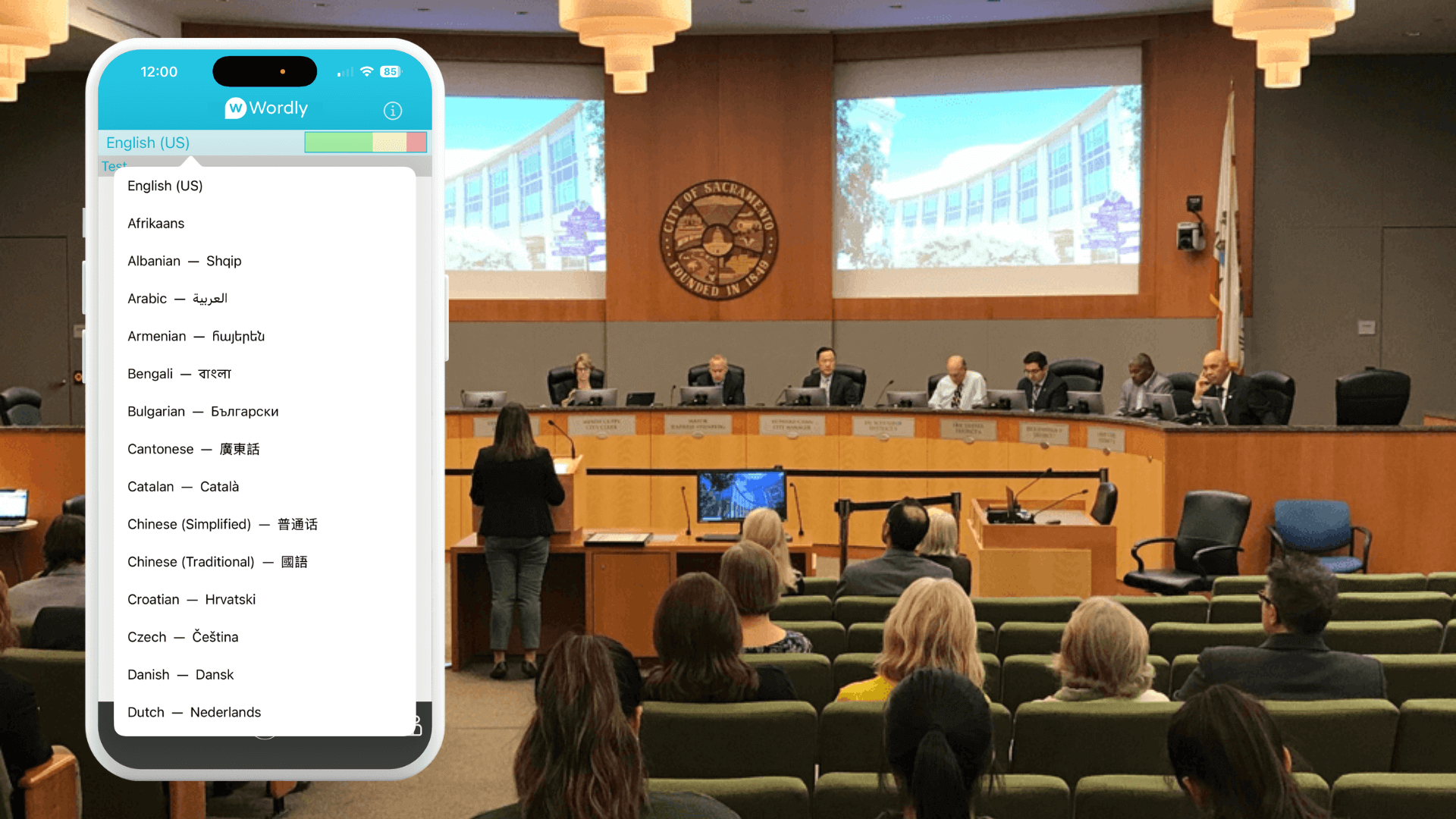 City council meeting in progress with attendees seated and a speaker addressing council members. A smartphone screen in the foreground displays Wordly