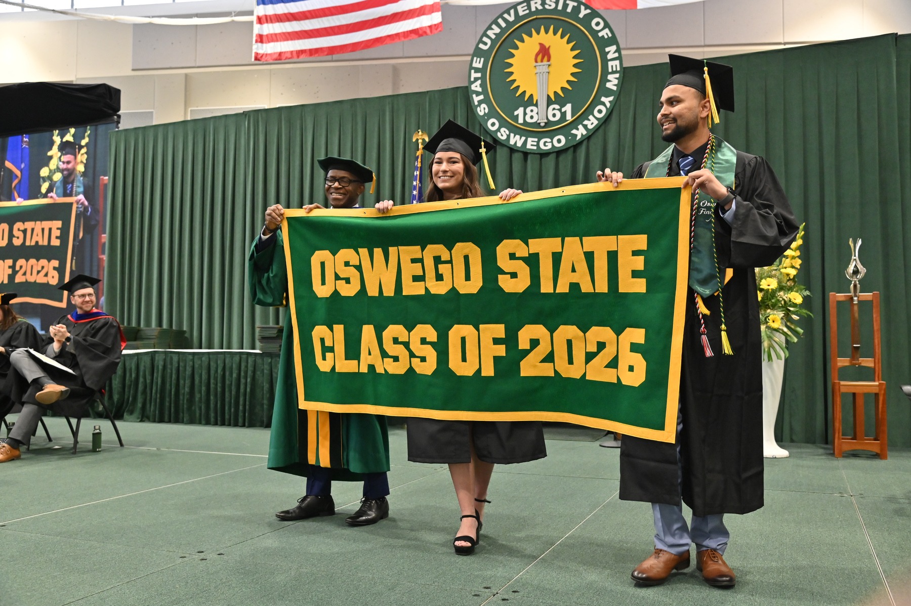 Graduates and a faculty member hold an Oswego State Class of 2026 banner on stage at SUNY Oswego’s May 2026 Commencement