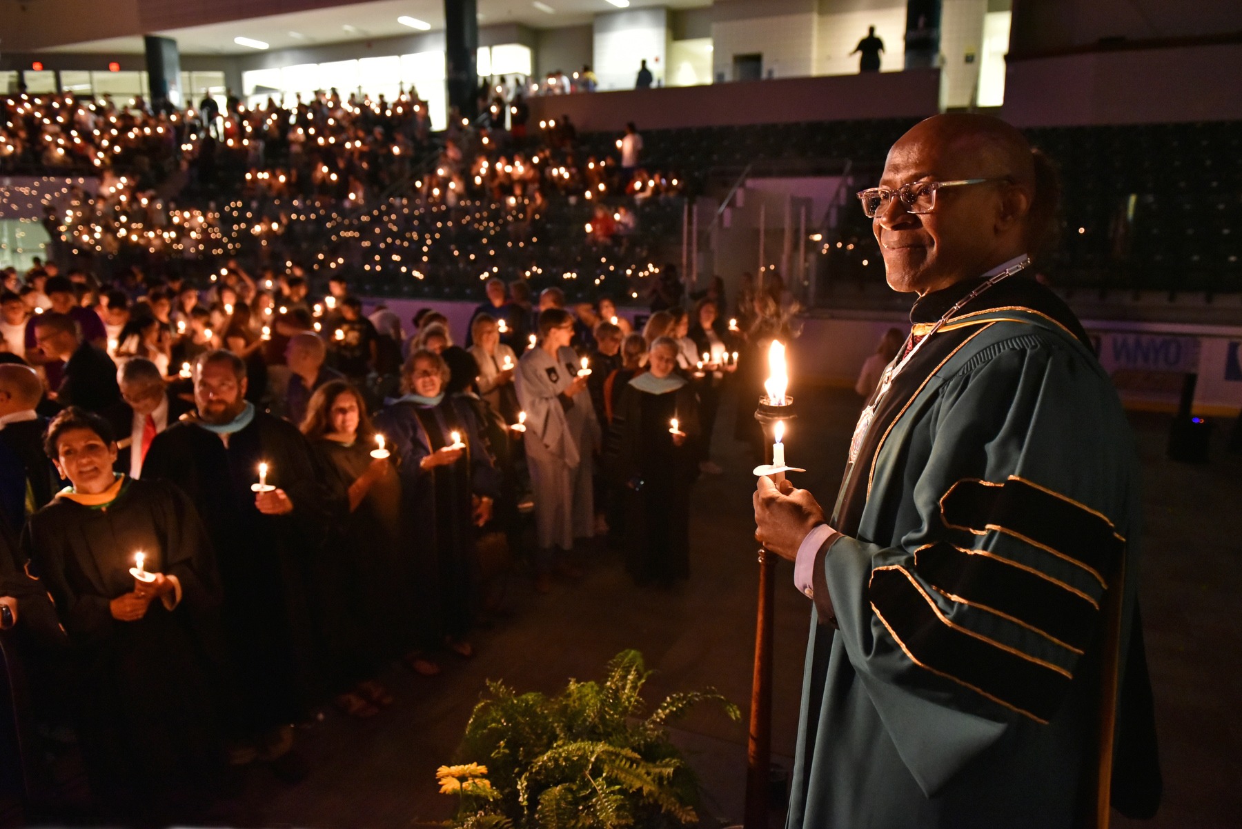 Faculty member holding a lit torch and candle at SUNY Oswego’s Torchlight ceremony, with faculty and new students holding candles in the arena