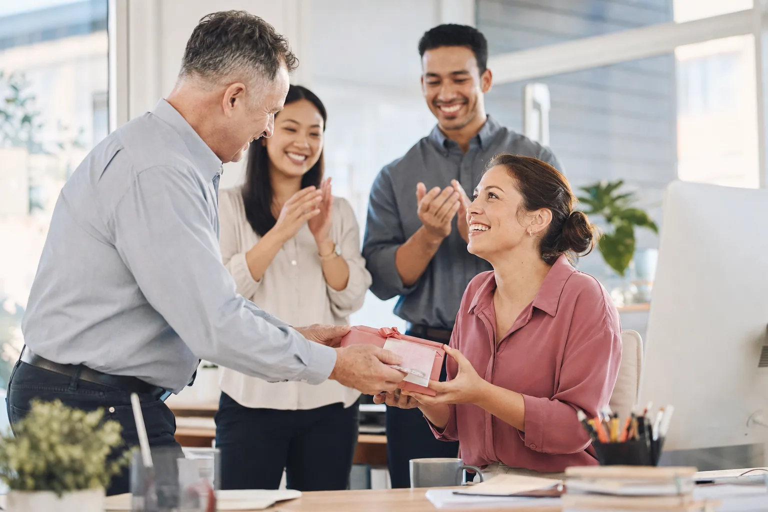 A manager handing a gift and card to a smiling employee while teammates clap