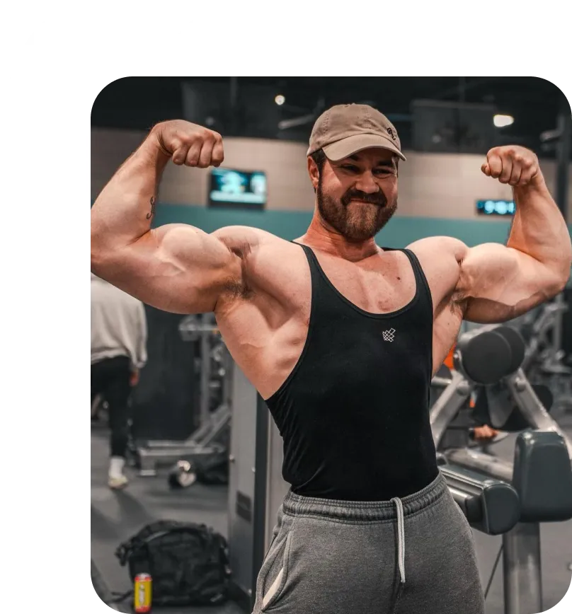 Man in a gym wearing a cap, black tank top, and gray sweatpants flexing his muscular biceps.
