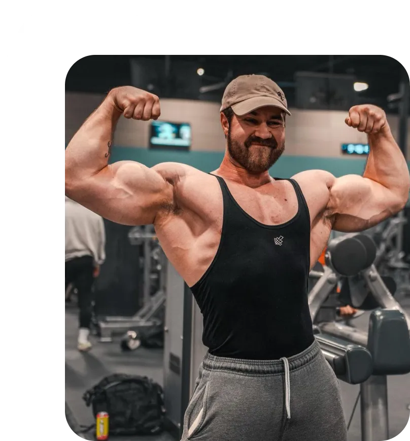 Muscular man in a black tank top and gray sweatpants flexing his biceps inside a gym.