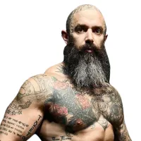Muscular man with a long gray beard, tattoos on chest and arms, and a bald head with a tattoo on the scalp.