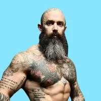 Muscular man with a long gray beard and tattoos covering his chest and arms against a blue background.