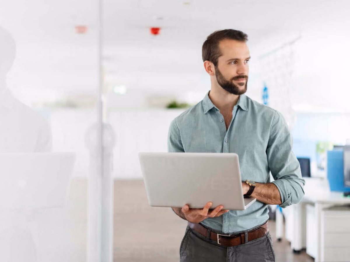 Bearded man in a green shirt holding a laptop and looking to the side in a modern office.