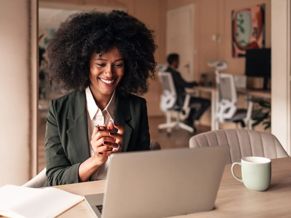 Smiling woman with curly hair sitting at a desk, looking at a laptop, with a man working in the background.