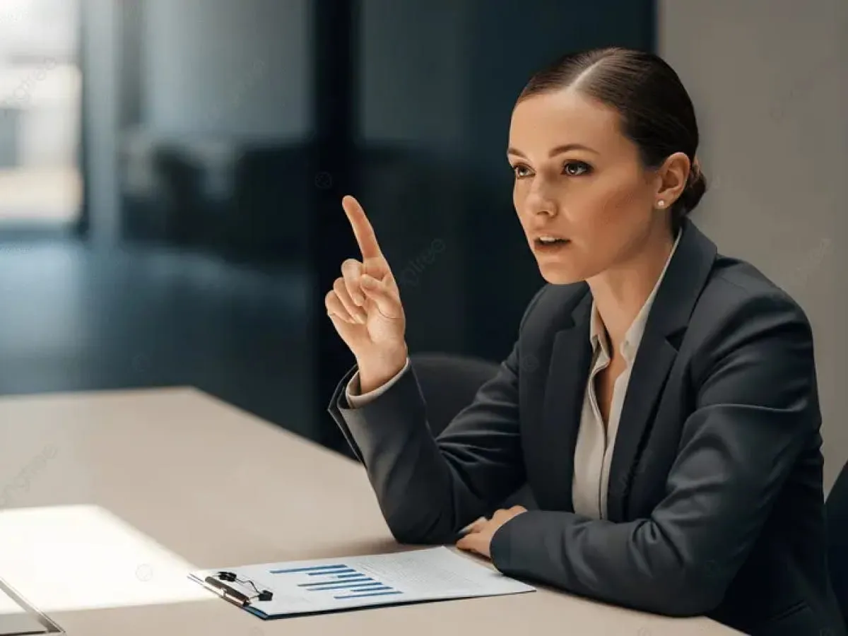 Businesswoman in a dark suit pointing upward while sitting at a table with documents and charts.