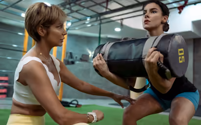 Two women exercising together, one holding a weighted sandbag while the other coaches her squat form in a gym.