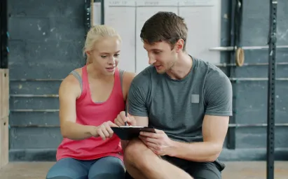 A woman in a pink tank top and a man in a gray t-shirt discuss a workout plan on a clipboard in a gym.