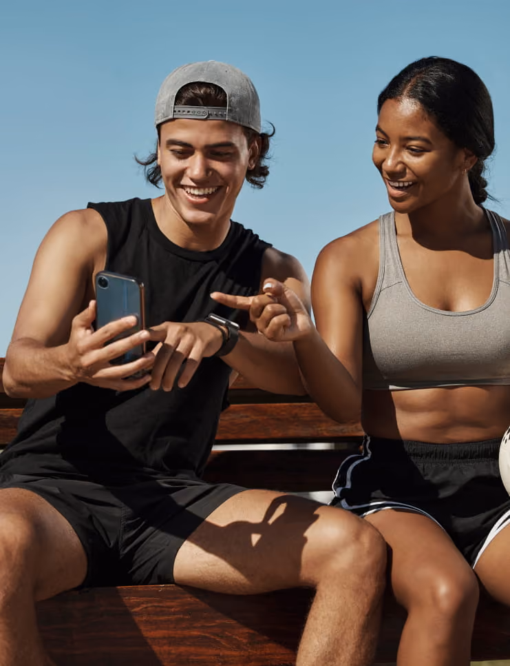 Smiling young man and woman sitting on a bench outdoors, looking at a smartphone together under clear blue sky.