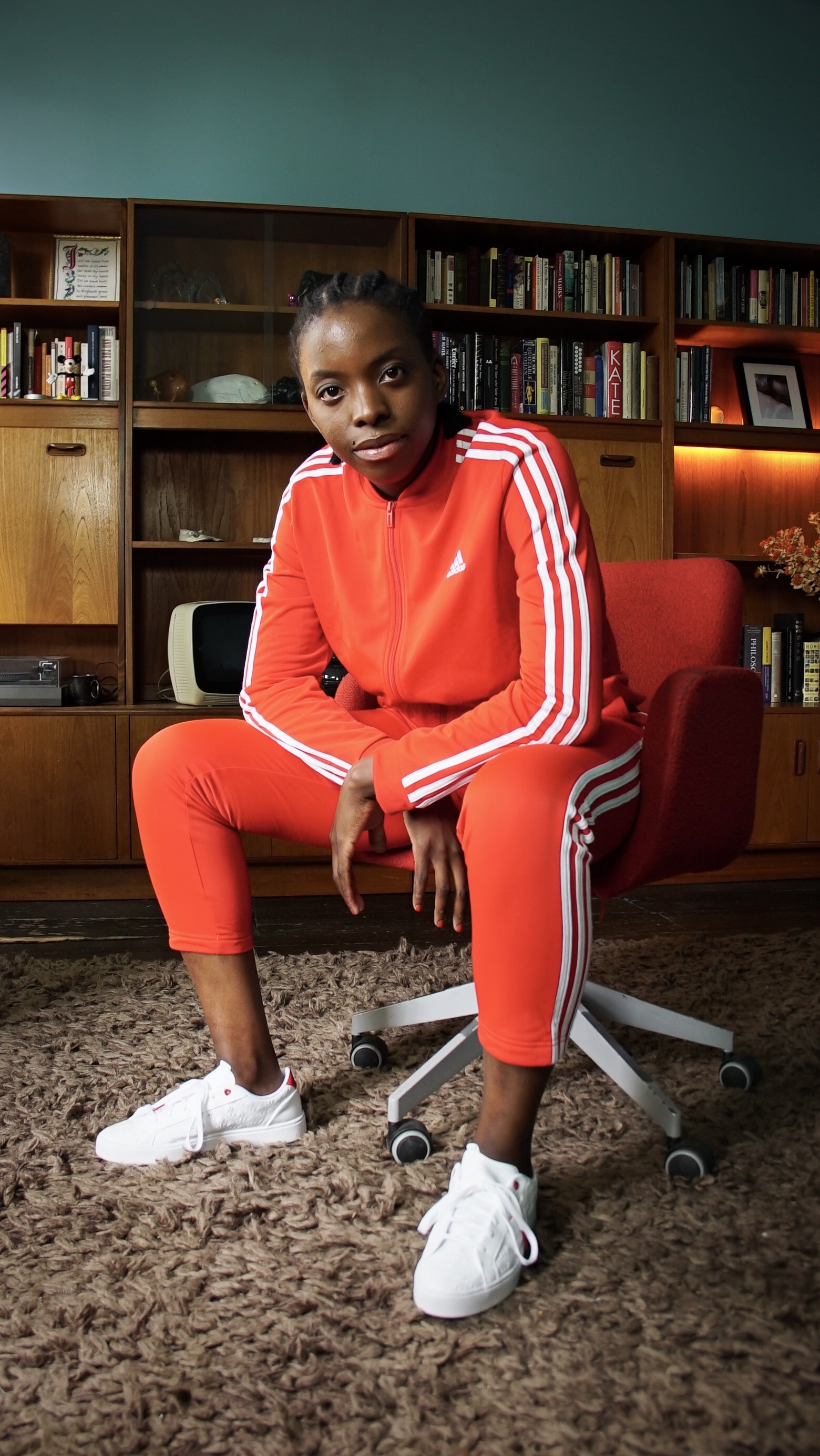 Foto di Vanessa Sauls in una libreria, seduta su una poltrona rossa e indossando una tuta Adidas rossa con strisce bianche.