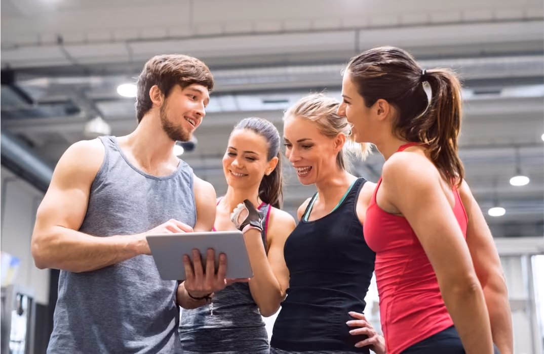 Group of fitness professionals evaluating personal trainer software pricing and features together on a digital tablet in a modern gym.