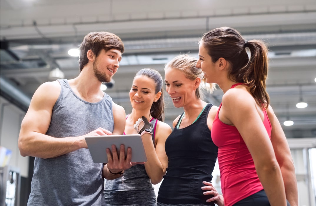 Group of fitness professionals evaluating personal trainer software pricing and features together on a digital tablet in a modern gym.