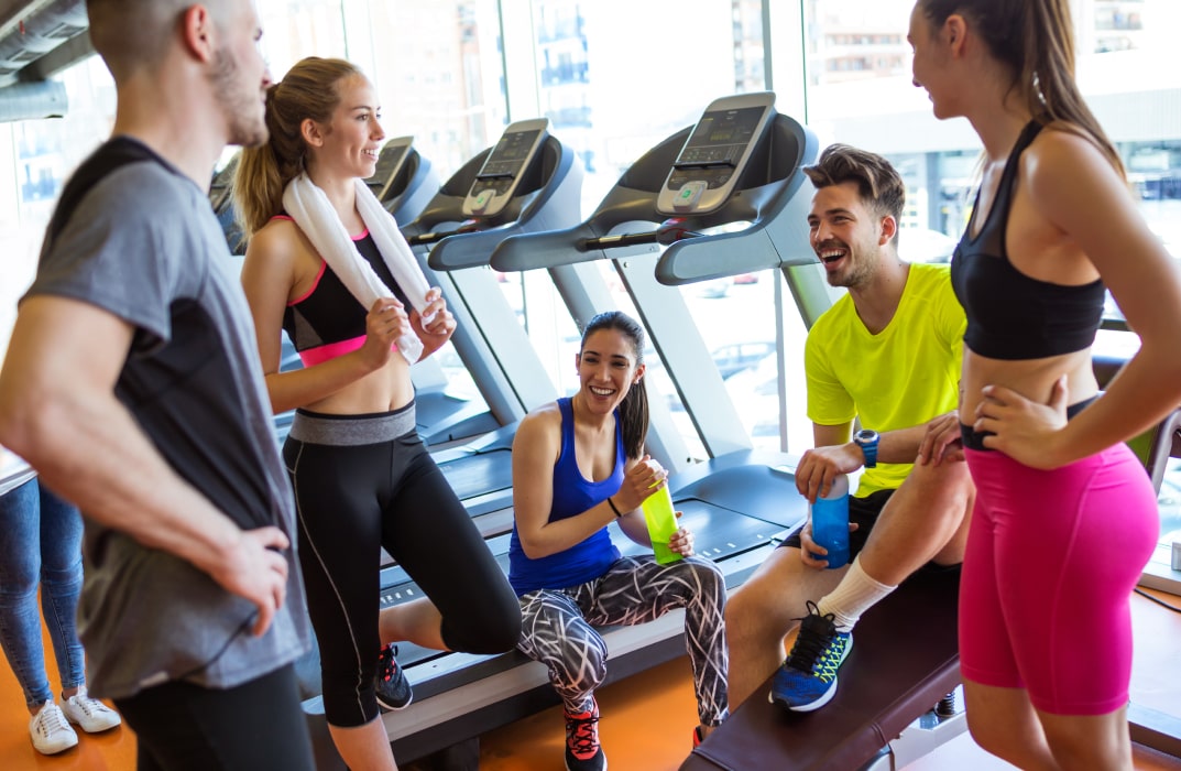 A group of participants is seen in a mirrored studio taking part in a lively fitness class, while a trainer works one-on-one with a client nearby.
